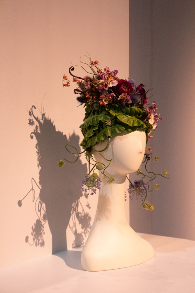 A white mannequin head with hat made of large green leaves and bouquet of flowers.