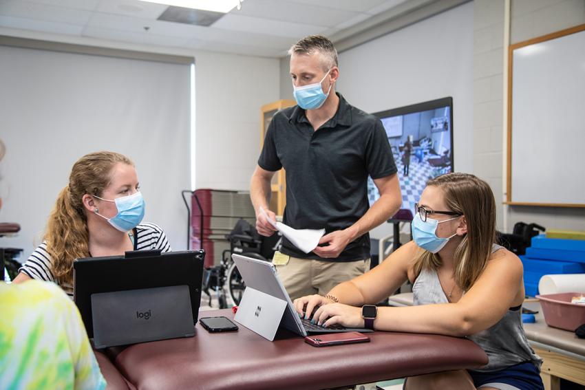 Dr. Michael Tevald works with two female students, all wearing masks.