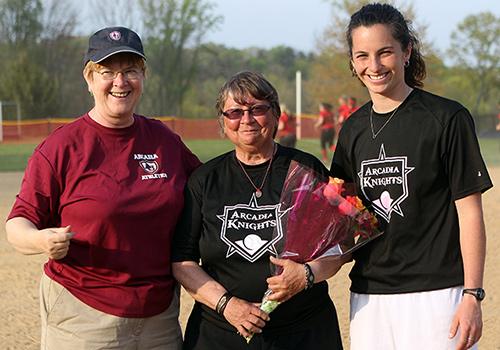 Three people wearing Arcadia Knights t-shirts smile, the middle person holding a bouquet of flowers
