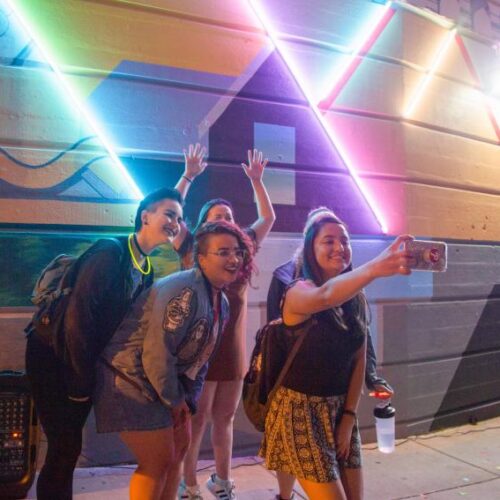 Young adults pose for a selfie at the Glenside Mural, lit with neon lights