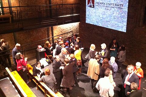 A group of several people socializing in a dark room with a projection that reads "ARCADIA UNIVERSITY"