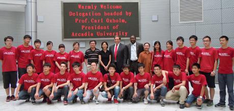 A large group of people wearing red, Arcadia shirts with 5 people in the center wear dress clothes with a sign behind them all