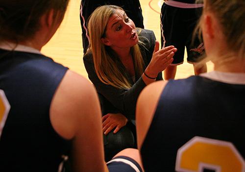 Coach Rose Katz crouches while gesturing and speaking to her basketball players around her