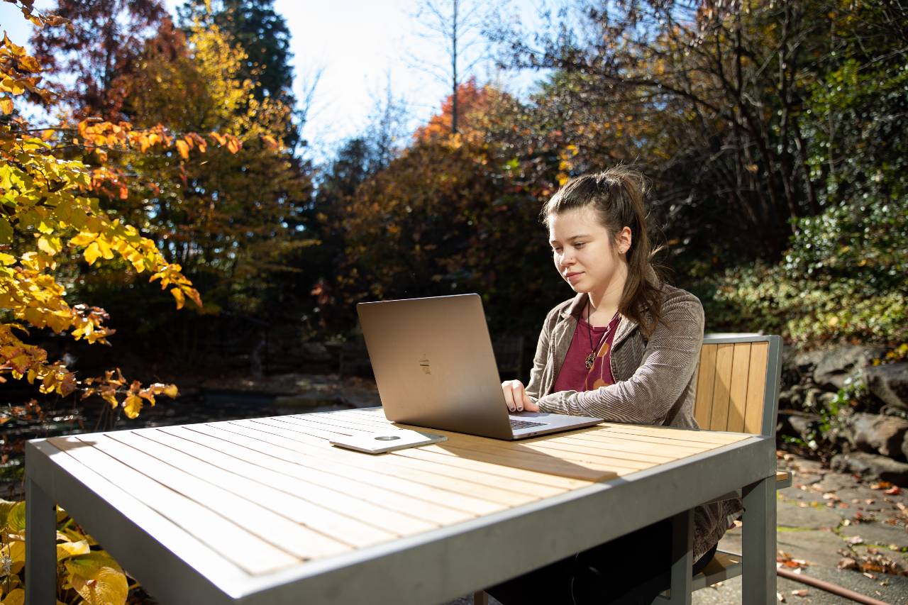 A student sitting outside during the day at a table, working on a laptop.