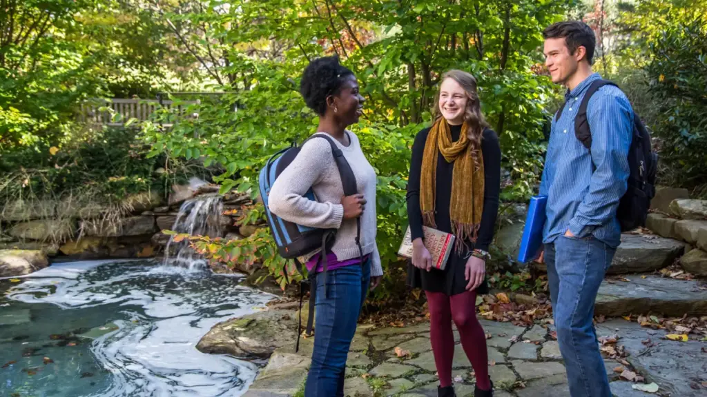 Smiling students stand next to Easton Pond wearing bookbags and carrying books