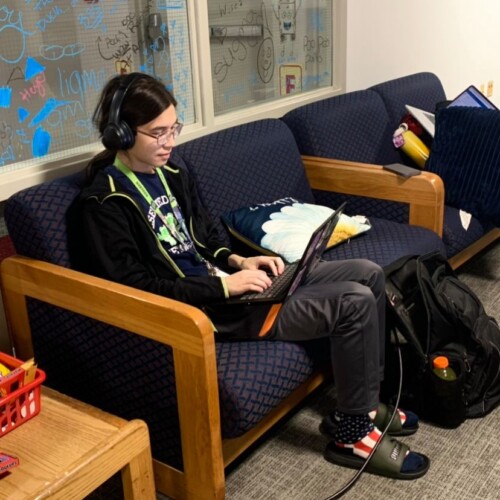 A student sits on a lounge couch wearing headphones and typing on a laptop
