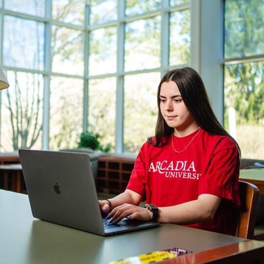 A female student in a red Arcadia shirt works at her laptop computer at a table surrounded by windows in Landman Library