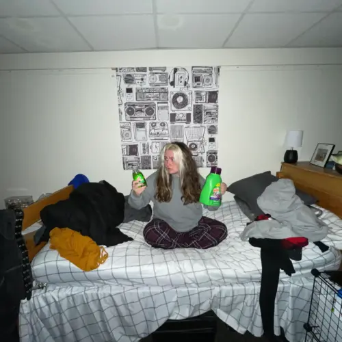 A student sits on a form room bed holding dish soap in one hand and laundry detergent in the other