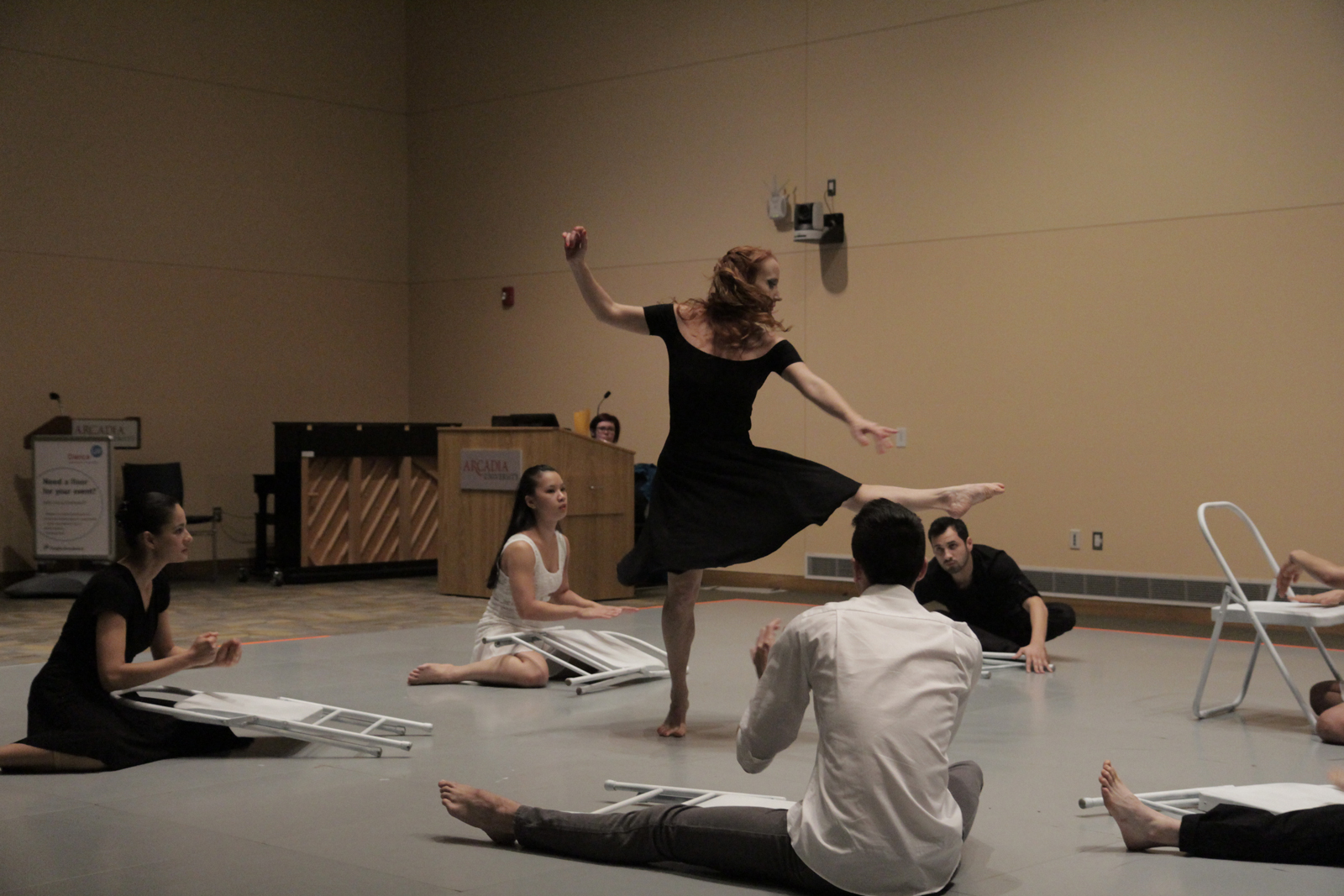 a group of people participating in a dance performance, one woman is standing with her left leg raised while six others are seated around her interacting with metal folding chairs