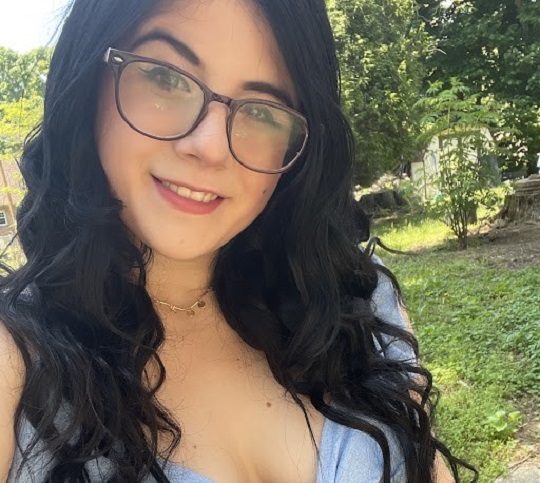 G.O.L.D. (graduate of the last decade) Alumna, Ashley Selig '19, poses outside for a self-taken photo. She has long, dark hair and glasses. She is smiling.