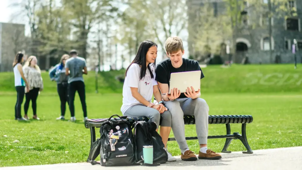 Two students share a look at a computer screen while sitting on the great lawn.