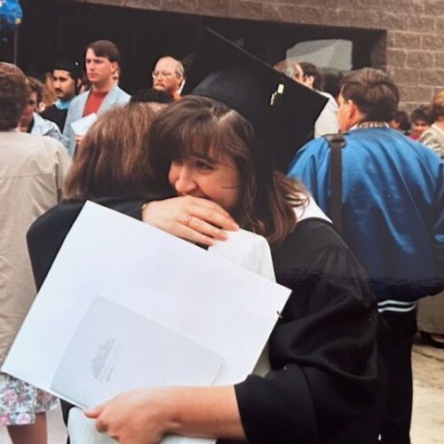 Winnifred and Sally are embracing in a hug. Sally is holding her degree in graduation regalia.