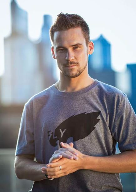 guy with brown hair and hands clasped in front of him. standing in front of Philly skyline in an Eagles gray shirt.