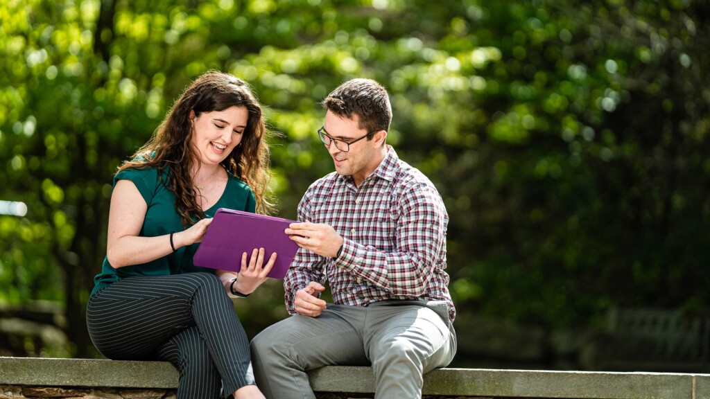 Two people sitting on a cement ledge looking a purple tablet