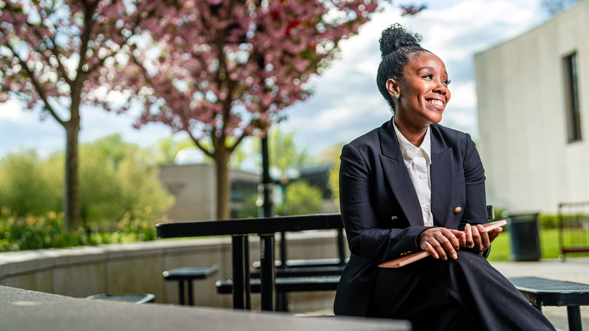 A Black woman in suit sitting down on benches while holding her laptop.