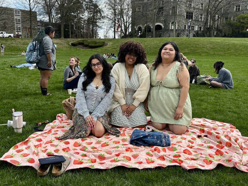 Three students wearing dresses sit on a strawberry picnic blanket on Haber Green