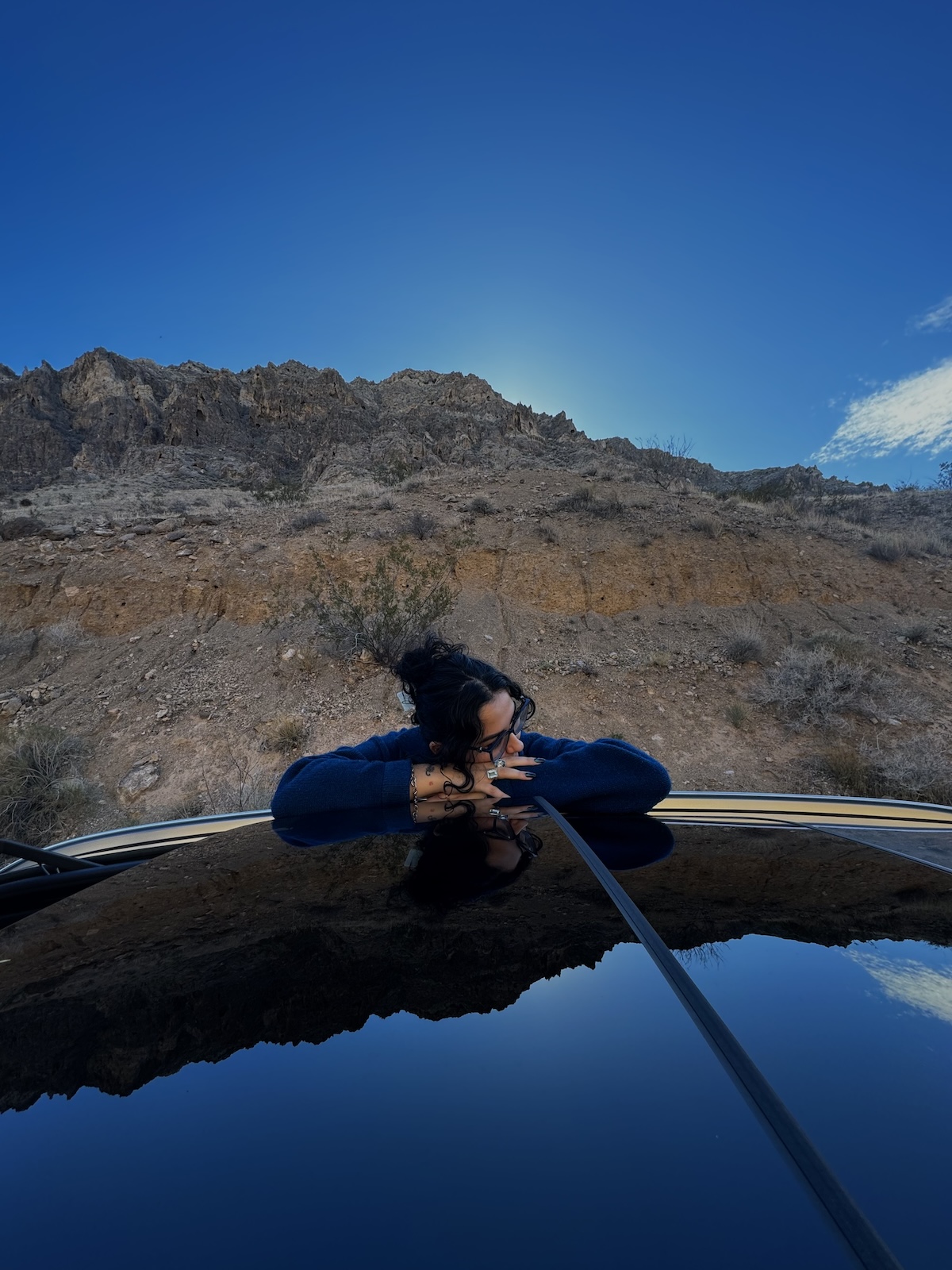 A woman rests her head on the roof of a car, with rocky mountain face behind her