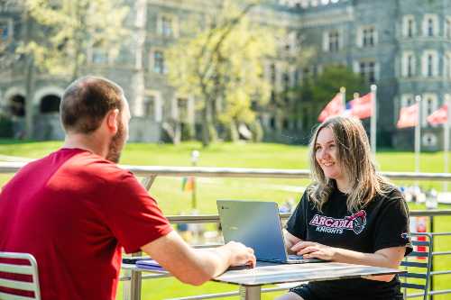 Two student sit opposite each other working on a laptop at an outdoor table in the commons