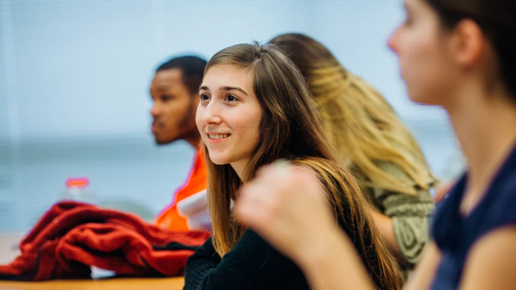 An undergraduate student smiles as she sits and leans forward during anundergraduate class lecture.