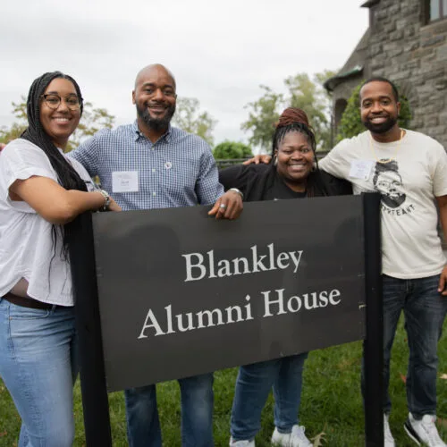 Alumni smiling at the camera and standing with the "Blankley Alumni House" sign.
