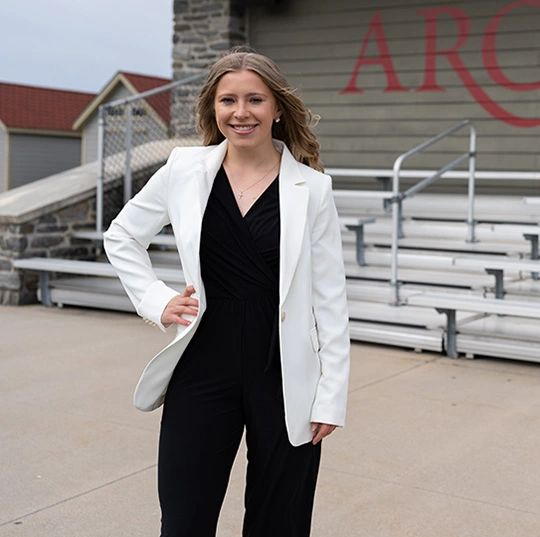 A sports management major wears a white suit and stands in an athletic field.