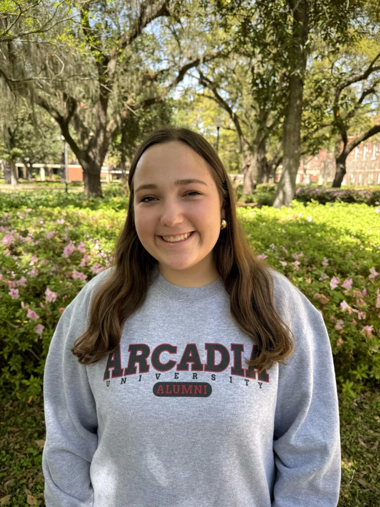 Anna is standing in front of green foliage wearing a gray Arcadia University crewneck sweatshirt.