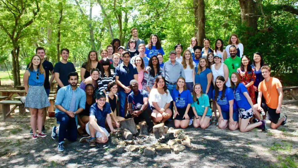 A large group of IPCR students site and stand by a firepit surrounded by big leafy trees overseas.