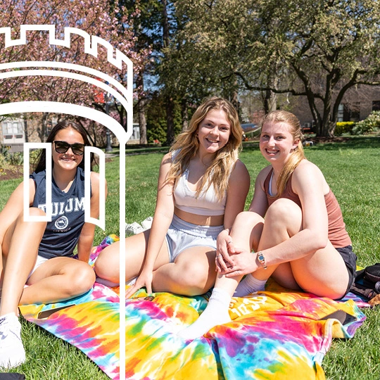 Three students sit on a beach blanket on the campus lawn.