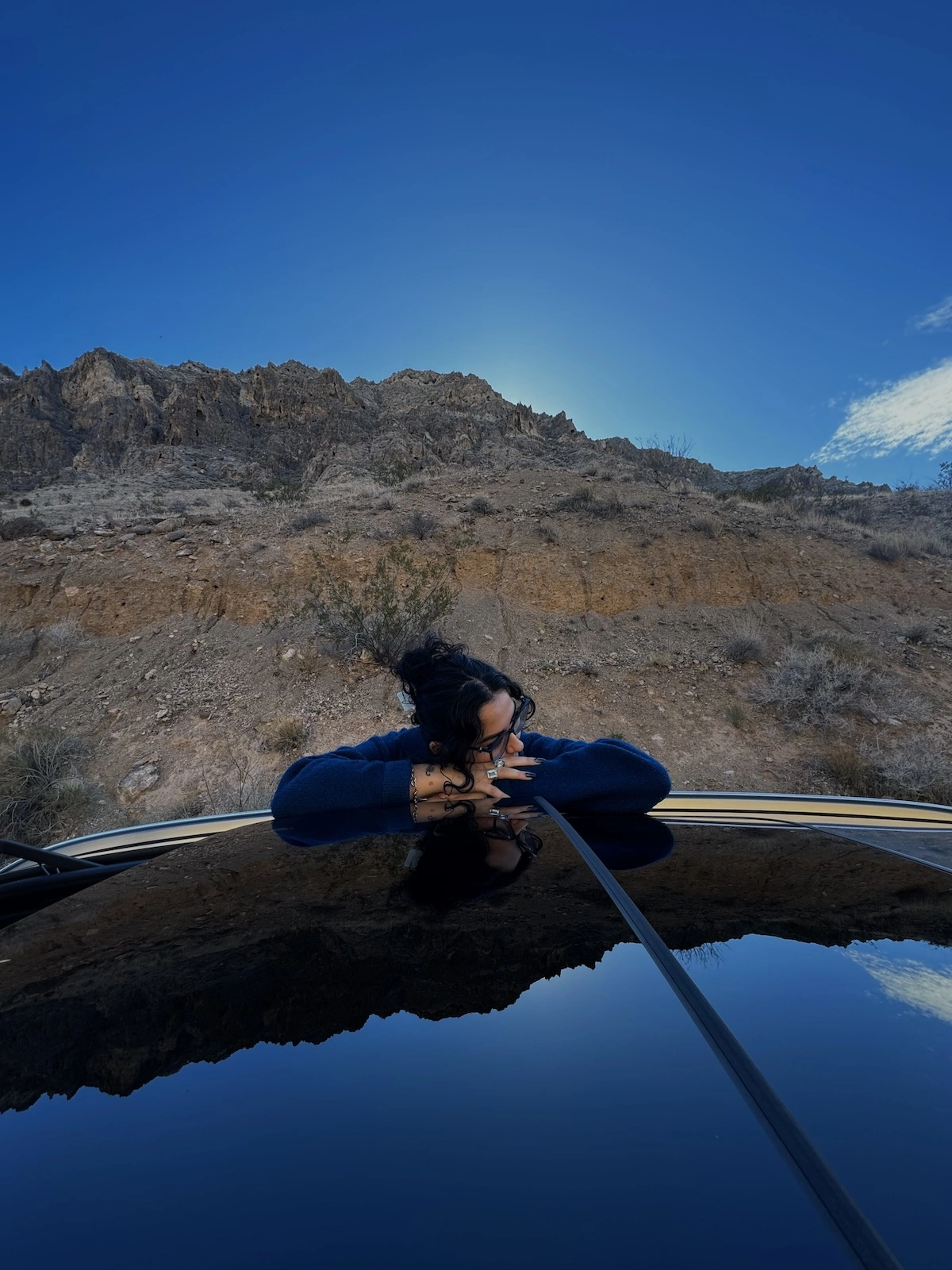 A woman rests her head on the roof of a car, with rocky mountain face behind her