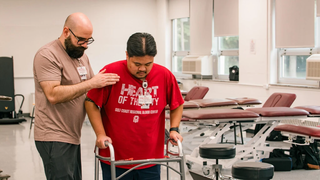 A physical therapy students works with a patient in a clinical setting lined with excercise tables and exam stools.