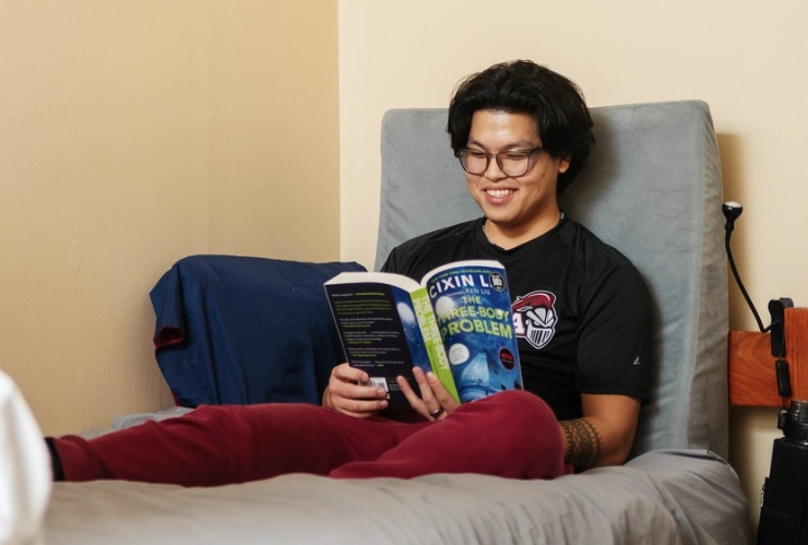 Lee Conlu '25, sitting on his dorm room bed and reading from a book.