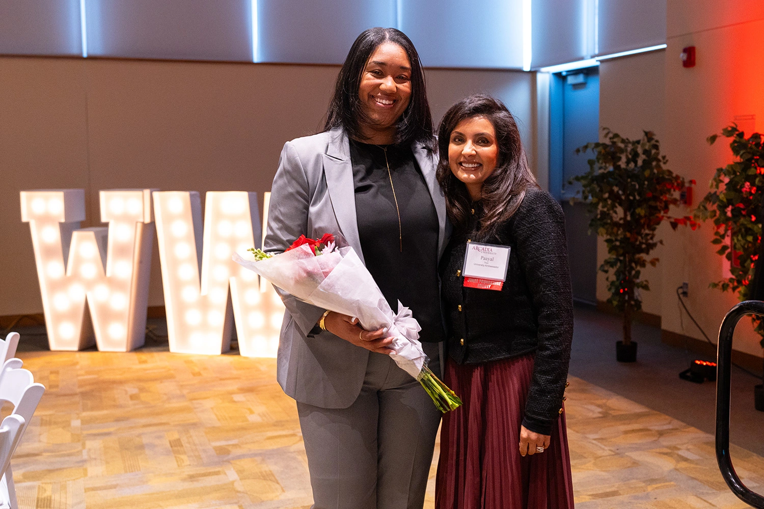 Tw women stand and one holds flowers in an event space decorated for the Women who Lead Forum.