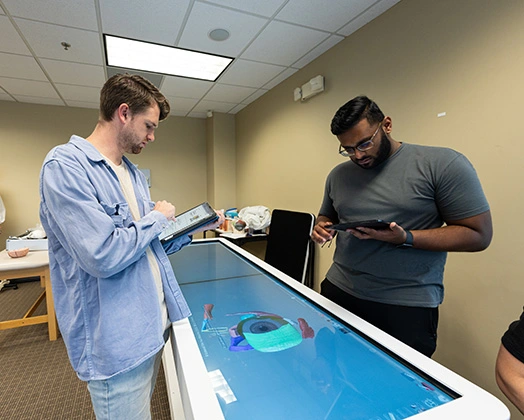 Two physician assistant students work on tablets over a digital exam table.