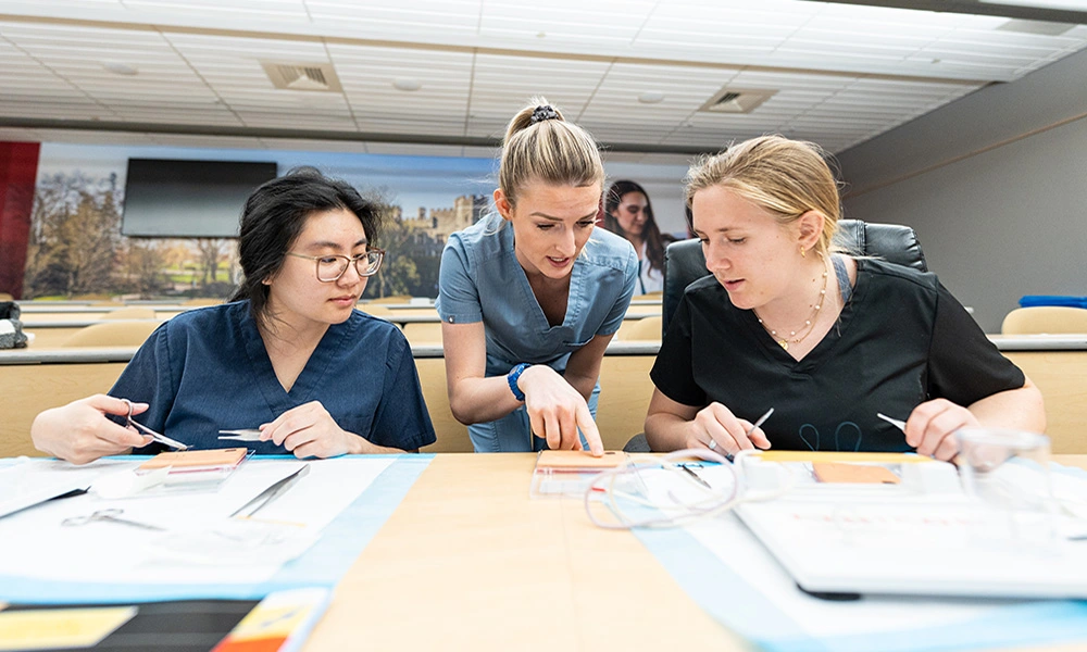 A physician assistant instructor points at a demo square to help two students during a classroom activity.