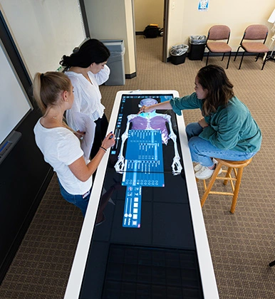 Three physician assistant students work over a digital exam table that has a digital skeleton.