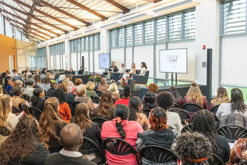 Women fill chairs on the second floor of University Commons facing a stage on which four women sit