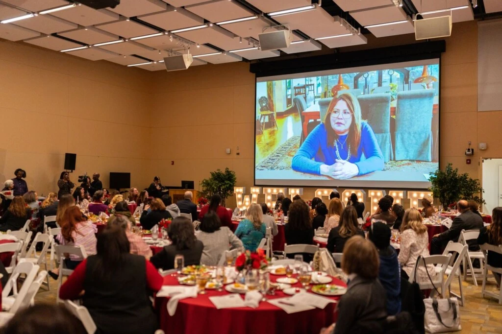 Women sit around red tableclothed tables staring at a large projector screen containing Rachael Ray